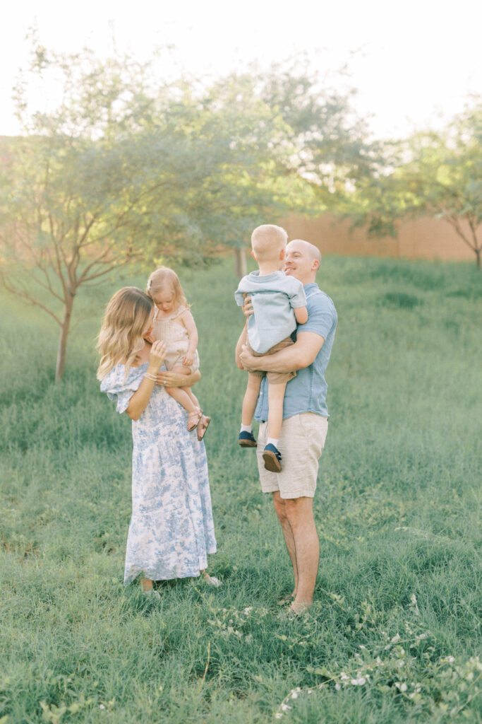 mom in blue dress holding little girl in tan romper standing in grass next her to husband holding their son.