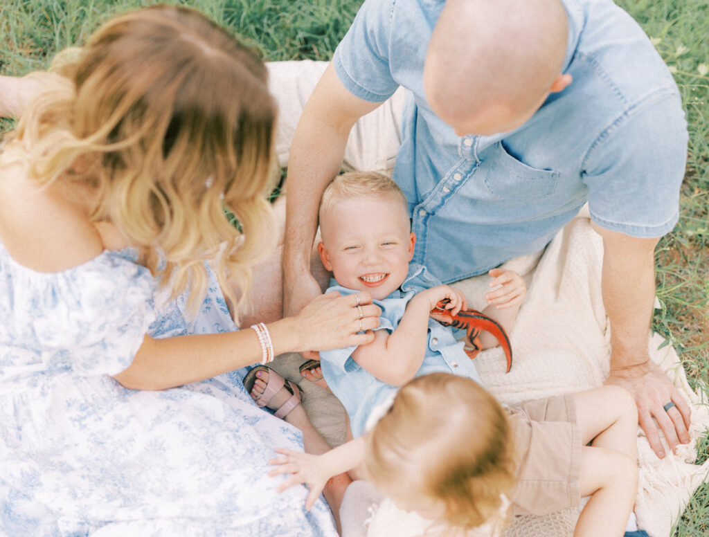 mom and dad sitting on ground tickling their son who is holding a red dinosaur during full-service family photography session