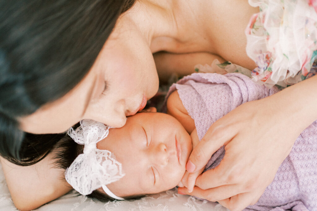 mom laying in bed tenderly kissing her newborn baby's cheek wrapped in a purple swaddle in a studio in Phoenix Arizona.