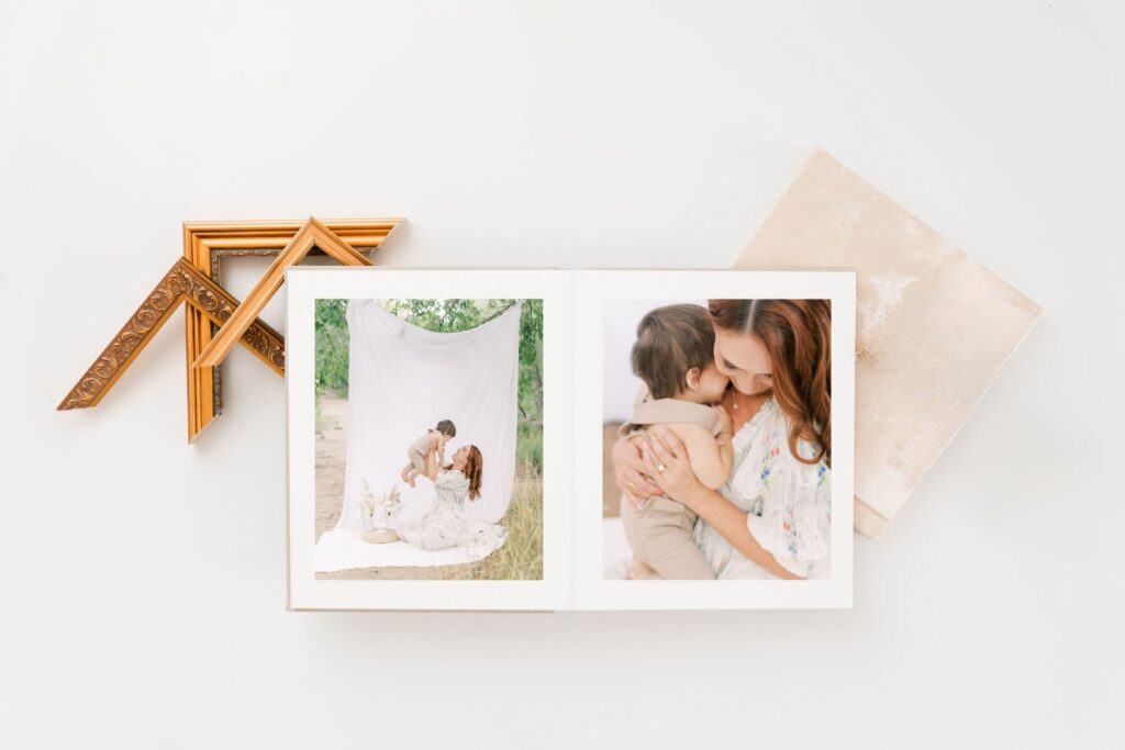 Image of an open Heirloom album with a photo of a mom holding up her one year old son while sitting on a white sheet with the white sheet pulled up in the background. The other image is a close up of mom in white floral dress snuggling her one year old son. Gold frames surround the album.