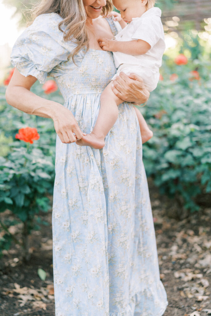 Close up of mom in blue dress holding toddler son in a rose garden playing with his foot in Scottsdale Arizona.