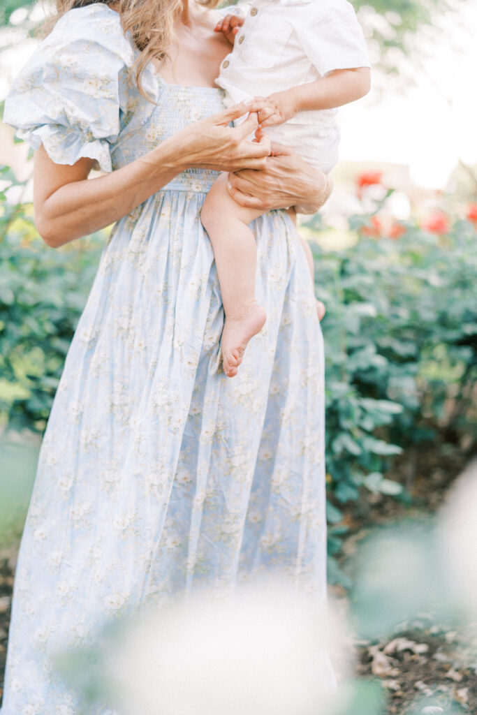 Mom in blue dress in a rose garden in Phoenix Az holding her toddler son and playing with his fingers.