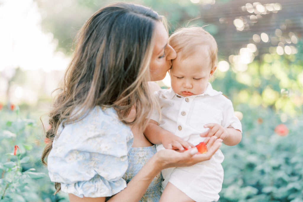 Close up photo of mom in a blue dress kissing toddler sons head while he looks at rose petals in a rose garden in Mesa Arizona.