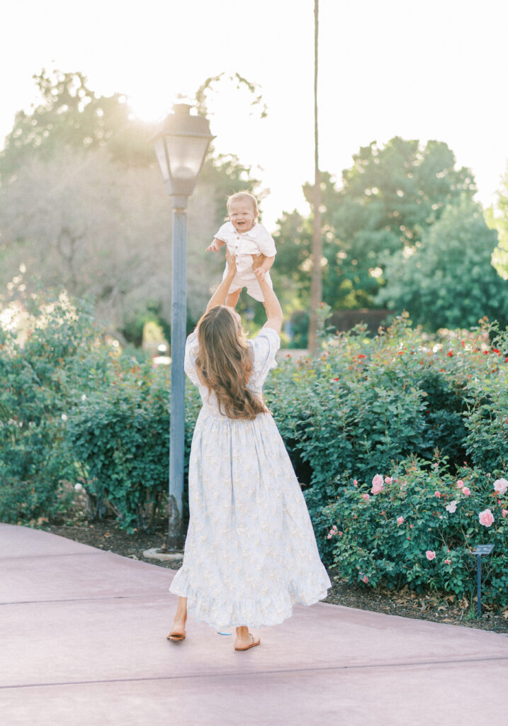 Mom in blue dress playing with toddler son, tossing his up in the air in a rose garden in Arizona. 