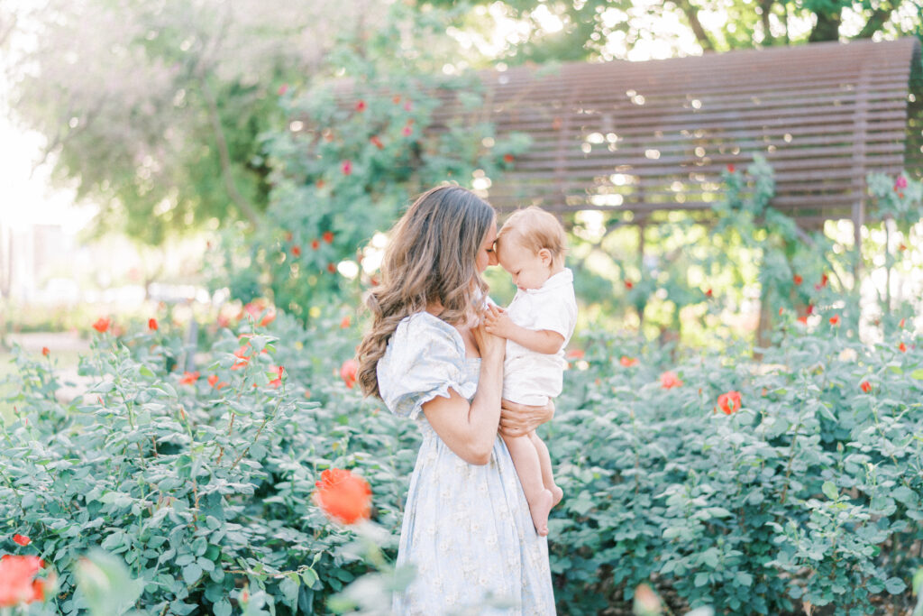 A Scottsdale Arizona mom in a light blue dress holding toddler son in white shirt among roses in a garden.