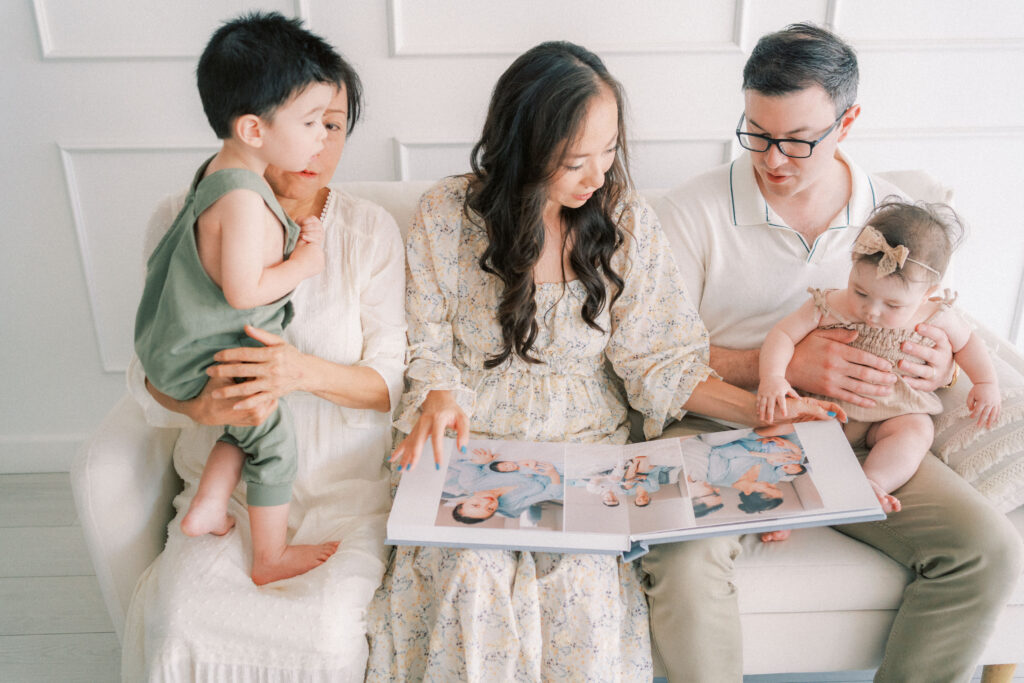 A mom, dad, grandma, toddler boy and 6 month baby girl looking through their heirloom album while sitting on a couch in a Mesa Newborn studio.