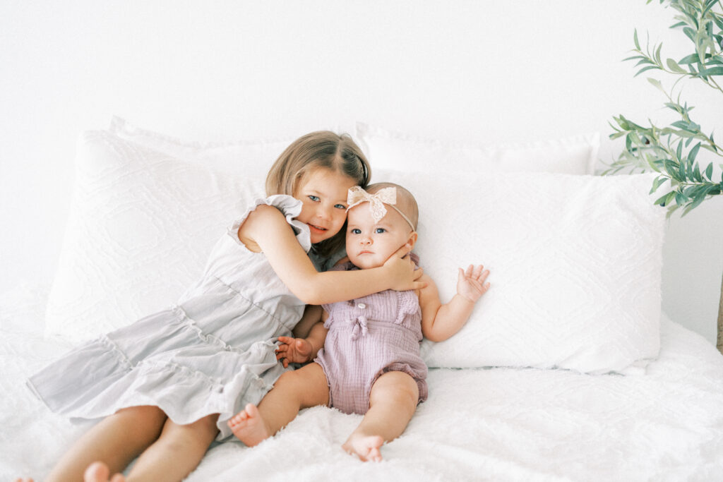 Toddler girl and 6 month old sister sitting on a bed in Scottsdale studio.