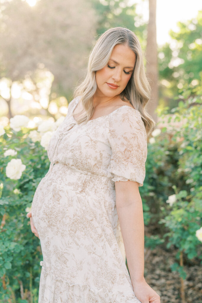 Pregnant woman in tan and white dress looking down in a field of roses in Scottsdale Arizona.
