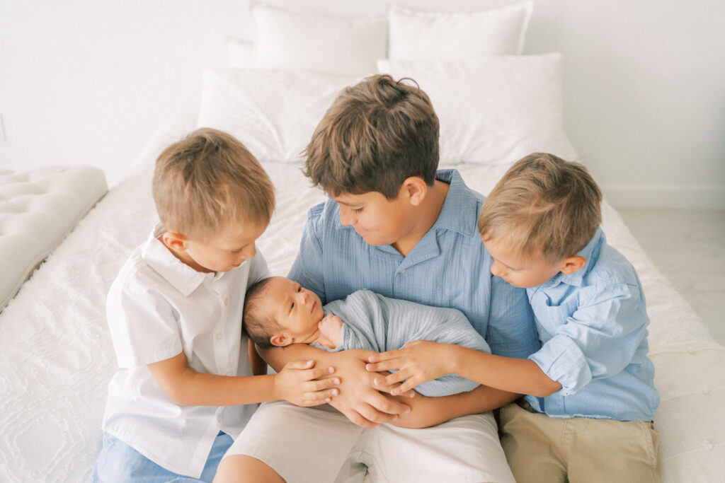 3 big brothers holding and looking down at baby newborn brother in white studio in Tempe Arizona